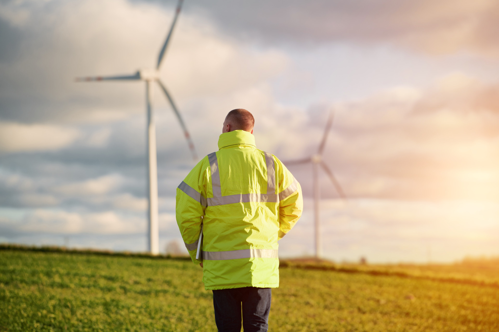 Mies keltaisessa työtakissa seisoo tuulivoimaloiden edessä, symboloiden uusiutuvaa energiaa ja kestävyyttä.